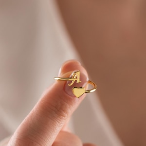 May include: A gold-colored ring with the letter "A" and a heart-shaped design. The ring is held up by a finger, showcasing its delicate details and elegant style. The ring is a piece of jewelry.