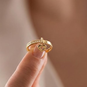May include: A gold ring with a heart-shaped charm. The ring band is adorned with small, clear stones. The heart charm hangs from a small bar. The ring is held up by a finger.