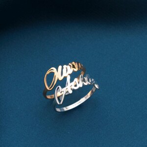 Puede incluir: Anillo personalizado con dos bandas, una dorada y otra plateada. La banda dorada presenta el nombre "Mary" en escritura cursiva, mientras que la banda plateada muestra "Ashli". El anillo se encuentra sobre una tela azul oscuro.
