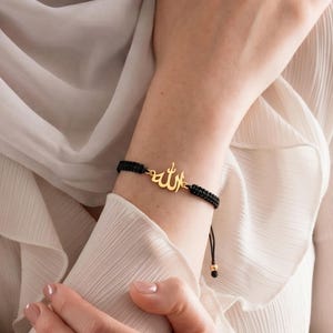 May include: A black cord bracelet featuring a gold-toned metal charm in the shape of the Arabic calligraphy for Allah. The bracelet has a woven texture and includes small gold-toned beads at the ends of the adjustable cord closure system.