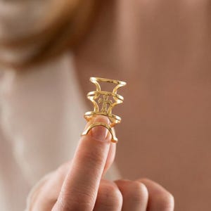 May include: A gold-colored ring with a cross design. The ring has a unique, open design with multiple bands and a central cross motif. The ring is held up by a finger.