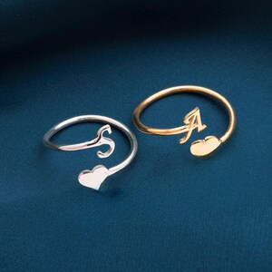 Könnte beinhalten: Zwei offene Ringe, einer silberfarben und einer goldfarben, jeweils mit einem kursiven Initialen und einem herzförmigen Akzent. Der silberne Ring hat den Buchstaben "S", und der goldene Ring hat den Buchstaben "A". Die Ringe werden auf einem dunkelblauen Hintergrund gezeigt.