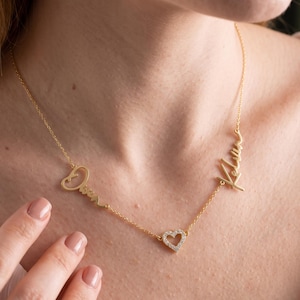 May include: A gold-toned necklace featuring a delicate chain with two cursive name pendants, Owen and Kelan, positioned on either side of a small, central heart-shaped charm encrusted with tiny clear stones that catch the light.