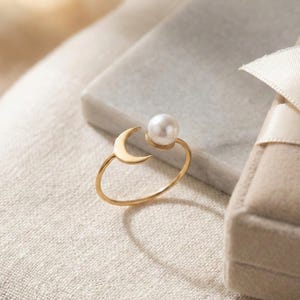 Puede incluir: Un anillo dorado con un diseño de media luna y una perla blanca. El anillo está sobre un fondo neutro, con un fondo borroso y una pequeña caja de regalo con una cinta crema. El diseño es delicado y elegante.