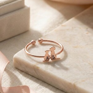 Peut inclure: Bague réglable en or rose avec deux silhouettes de chiens et les lettres "A" et "S". La bague est posée sur une surface en marbre, avec un écrin en velours et un ruban rose en arrière-plan. Un bijou unique.