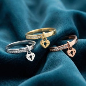 May include: Three rings with heart-shaped lock pendants. One ring is gold with a row of small clear stones, another is silver with clear stones, and the third is rose gold with clear stones. All have a heart-shaped lock pendant.