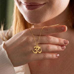 May include: A gold-coloured necklace featuring an Om symbol pendant. The pendant is a stylized representation of the sacred Hindu symbol. The necklace is held in a person's hand, showcasing the delicate chain and pendant design.