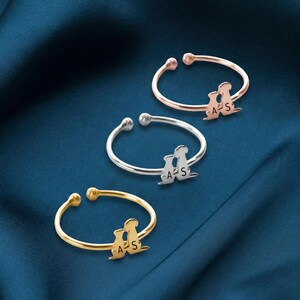 Peut inclure: Trois bagues ouvertes en or, argent et or rose. Chaque bague présente un motif de silhouette avec les lettres "A" et "S". Les bagues sont présentées sur une surface bleu foncé texturée.