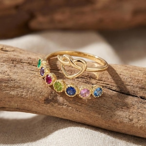 Puede incluir: Un anillo de oro con un diseño en forma de corazón y una banda adornada con piedras preciosas de colores. El anillo está colocado sobre un trozo de madera, mostrando su elegante diseño y colores vibrantes.