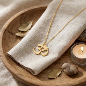 May include: A gold-colored necklace with an Om symbol pendant rests on a beige linen cloth. The necklace is displayed on a wooden tray with a lit candle and decorative leaves. The Om symbol is a spiritual icon.