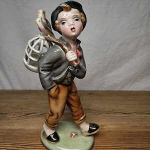 Könnte beinhalten: Eine Porzellanfigur einer Person mit dunkelgrüner Mütze, grauer Jacke und beigefarbener Hose. Die Figur trägt einen Holzstock über der Schulter, an dessen Ende ein weißer Vogelkäfig mit zwei kleinen Vögeln befestigt ist, auf einem Sockel.
