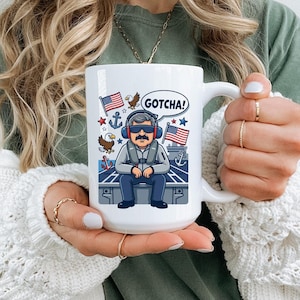 May include: White ceramic mug featuring a cartoon illustration of a man in a pilot's uniform with the text "GOTCHA!". The design includes American flags, eagles, and stars. The mug has a large handle.