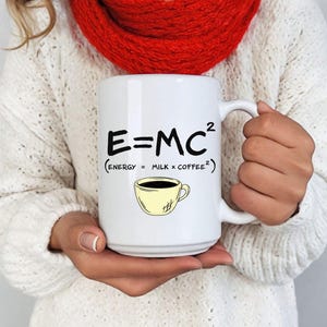 May include: White ceramic coffee mug with the equation E=MC² and the text "(Energy = Milk x Coffee²)". A yellow coffee cup graphic is below the text. The mug is held by a person wearing a white sweater and a red scarf.