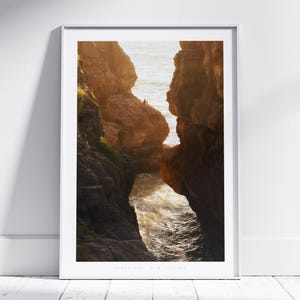 Puede incluir: Una impresión enmarcada de un paisaje costero con imponentes formaciones rocosas y el océano. La imagen captura un arco natural con la luz del sol iluminando el agua. La impresión está en un marco blanco y se muestra en una pared blanca.