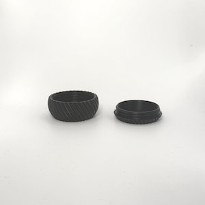 Peut inclure: Deux récipients circulaires noirs avec un motif texturé et strié. Un récipient est ouvert, révélant l'intérieur. Les récipients sont faits d'un matériau sombre et reposent sur une surface blanche.