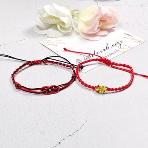 Può includere: Due braccialetti di corda rossa con un design a nodo. Un braccialetto ha una corda nera intrecciata, e l'altro ha un ciondolo a forma di fiore dorato.