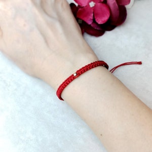 Puede incluir: Una pulsera roja de macramé con una pequeña cuenta dorada. La pulsera está anudada y tiene lazos ajustables. La pulsera se lleva en la muñeca.