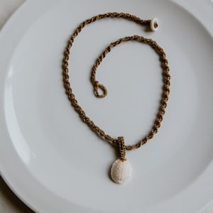 Puede incluir: Un collar artesanal con un cordón trenzado marrón. Presenta un colgante ovalado blanco con una superficie texturizada. El collar se exhibe sobre un plato blanco.