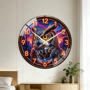 May include: A round wall clock featuring a detailed owl illustration with a desert sunset backdrop. The clock has a brown frame, gold hands, and neon orange numerals. The design includes a blue and red neon emblem at the bottom.