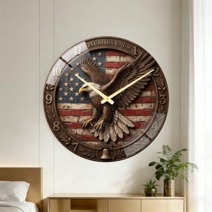 May include: A round wall clock featuring an American flag design with a detailed eagle in the center. The clock has gold-colored hands and a brown frame with the words "PLURIBUS UNUM" at the top. The clock is mounted on a light-colored wall.