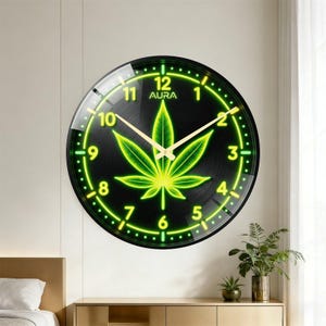 Pode incluir: Um relógio de parede redondo com mostrador preto e um gráfico de folha de canábis em verde néon brilhante no centro. O relógio apresenta números verdes luminosos de 1 a 12, ponteiros finos de cor dourada e a palavra AURA impressa em branco.
