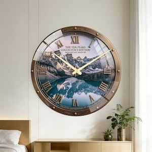 Op de afbeelding: Een ronde wandklok met een bronzen metalen frame en een glazen wijzerplaat. De wijzerplaat toont een landschap van een bergmeer en Romeinse cijfers als uurmarkeringen. De tekst "THE TEN PEAKS COLLECTOR'S EDITION" is zichtbaar.