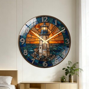 May include: Round wall clock with a stained glass design featuring a lighthouse scene. The clock face has a brown metal frame, gold hands, and large, silver-colored numerals. The lighthouse is white with a blue top, set against an orange and blue background.