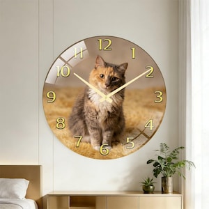 May include: A round wall clock featuring a cat image. The clock face has gold-colored numerals and hands. The cat has a mix of brown, orange, and white fur. The clock is set against a light brown background.