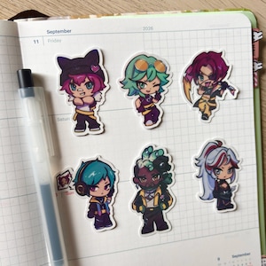 May include: Six die-cut vinyl stickers featuring stylized anime-inspired characters with vibrant hair colors and distinct outfits, arranged on a grid-lined planner page next to a black and white retractable gel pen. The characters display various expressions.