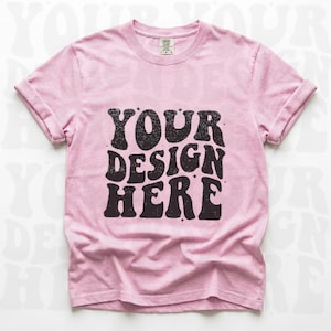 Könnte beinhalten: Hellrosa T-Shirt mit hochgekrempelten Ärmeln. Auf der Vorderseite steht der Text "YOUR DESIGN HERE" in schwarzer Retro-Schrift. Das T-Shirt hat einen Rundhalsausschnitt und ein Etikett am Halsausschnitt.