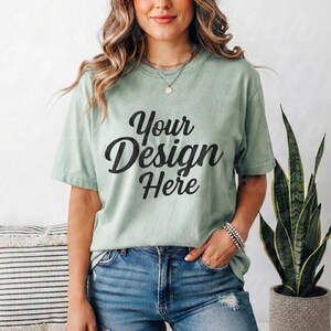 May include: A pale green t-shirt featuring the text "Your Design Here" in black script. The model is wearing blue denim jeans and a silver necklace. The shirt is a crew neck with short sleeves.