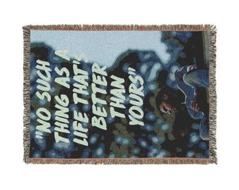 Puede incluir: Manta tejida con borde de flecos. La manta presenta un fondo azul y verde con el texto "NO SUCH THING AS A LIFE THAT'S BETTER THAN YOURS" en blanco. Una persona es parcialmente visible en el lado derecho.