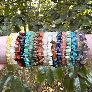 May include: A selection of gemstone chip bracelets in a variety of colours, including yellow, red, turquoise, pink, blue, and purple. The bracelets are displayed on a pink cylindrical surface against green foliage.