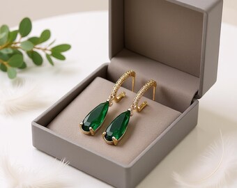 May include: A pair of gold-colored hoop earrings with teardrop-shaped, emerald-green gemstones, displayed in an open gray jewelry box. The earrings have a textured design on the hoops. The box is lined with a light brown fabric.