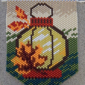 May include: A beaded wall hanging featuring a stylized image of a lemon with a red and brown rimmed jar. The lemon is yellow with white highlights, and the background is green, orange, and beige. The banner is made of small, colorful beads.
