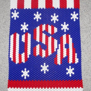 May include: A beaded wall hanging with a patriotic theme. The design features the letters "USA" in red and white stripes against a blue background with white star accents. Red and white stripes border the top and bottom.