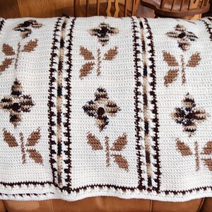 May include: Cream-colored crocheted blanket with a repeating pattern of brown flowers and butterflies. The blanket is rectangular and has a decorative border. The design is symmetrical and the blanket is folded over a brown surface.