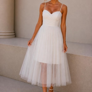 Puede incluir: Un vestido blanco de largo midi con escote corazón, tirantes finos y un corpiño ajustado con diseño plisado. La falda consta de una capa interior opaca cubierta por una capa superpuesta de tul transparente y voluminoso que cae debajo.