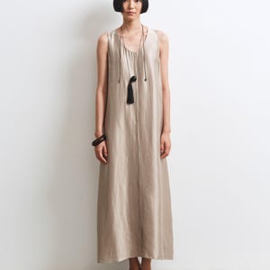 May include: A sleeveless, floor-length beige dress with a scoop neckline and a vertical seam down the center. The outfit includes a long, thin black cord necklace with a dark spherical pendant and tassel, a dark bangle, and black mesh flats.