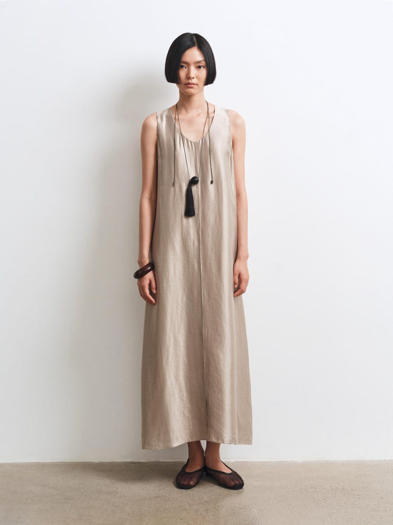 May include: A sleeveless, floor-length beige dress with a scoop neckline and a vertical seam down the center. The outfit includes a long, thin black cord necklace with a dark spherical pendant and tassel, a dark bangle, and black mesh flats.