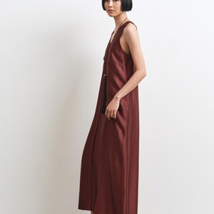 May include: A sleeveless, floor-length dress in a deep, rich burgundy hue. The garment features a straight, relaxed silhouette with a V-neckline and a vertical row of dark, circular buttons running down the front. The fabric has a subtle, natural sheen.
