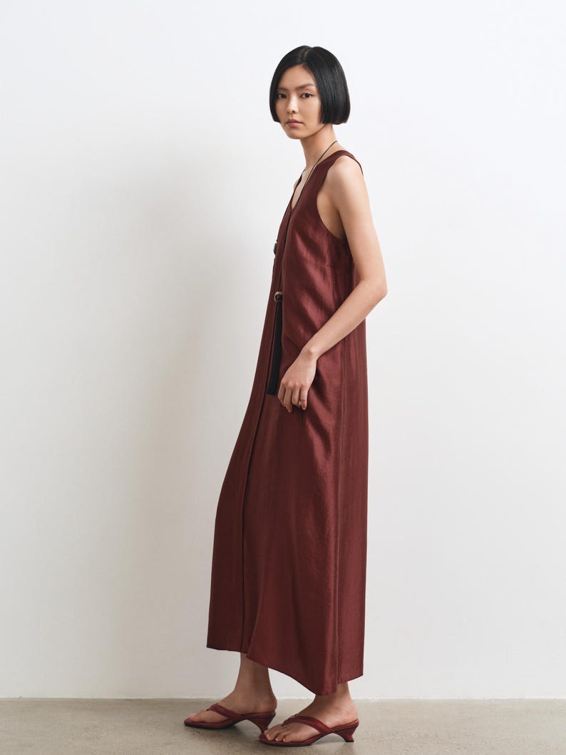 May include: A sleeveless, floor-length dress in a deep, rich burgundy hue. The garment features a straight, relaxed silhouette with a V-neckline and a vertical row of dark, circular buttons running down the front. The fabric has a subtle, natural sheen.