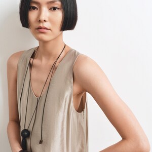 May include: A sleeveless beige top with a V-neckline, paired with a long black cord necklace featuring a large round black pendant. A thick, dark brown polished bangle is worn on the wrist. The garment has a smooth, slightly lustrous finish.