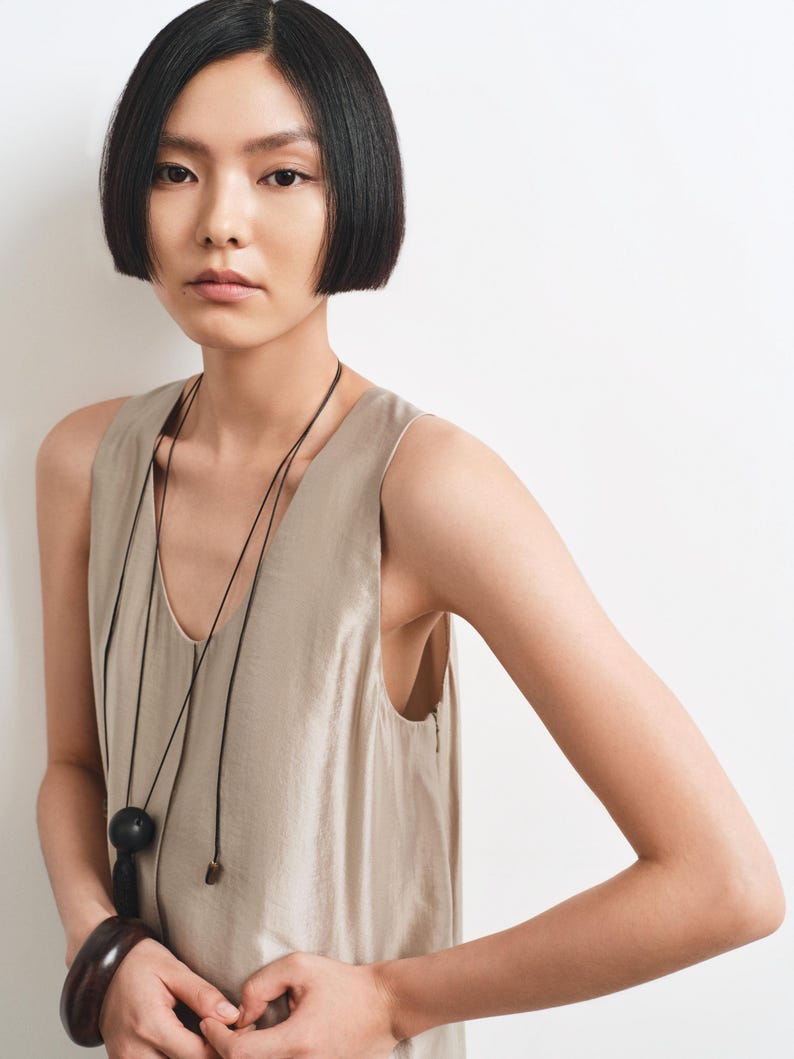May include: A sleeveless beige top with a V-neckline, paired with a long black cord necklace featuring a large round black pendant. A thick, dark brown polished bangle is worn on the wrist. The garment has a smooth, slightly lustrous finish.