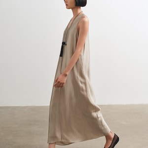 May include: A sleeveless, floor-length dress in a light taupe, satin-like fabric with a loose, flowing silhouette. The dress is paired with a long, thin black cord necklace featuring a black tassel pendant and sheer black mesh ballet flats.