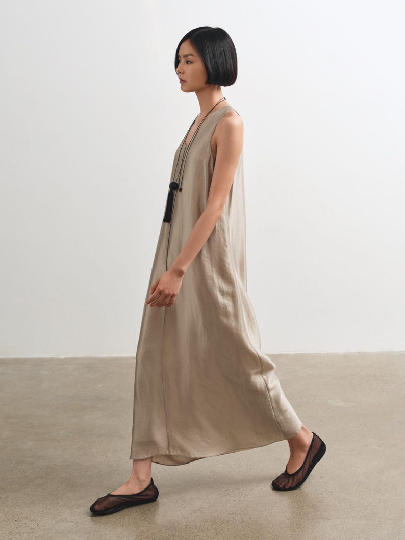 May include: A sleeveless, floor-length dress in a light taupe, satin-like fabric with a loose, flowing silhouette. The dress is paired with a long, thin black cord necklace featuring a black tassel pendant and sheer black mesh ballet flats.
