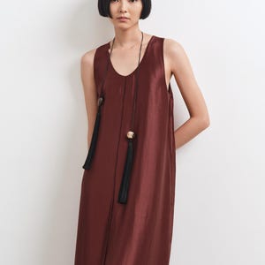 May include: A sleeveless, floor-length dress in a deep burgundy, satin-like fabric with a subtle sheen. The dress features a round neckline and a long, thin black cord necklace adorned with two decorative beads and two long black tassels.