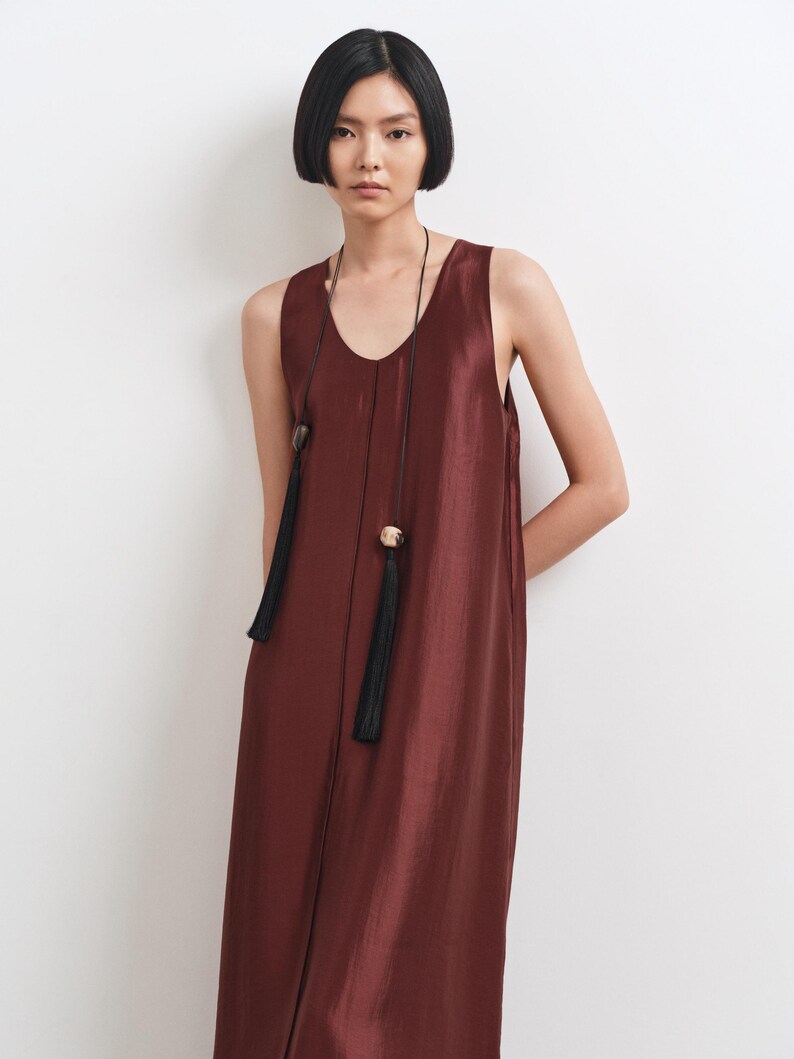 May include: A sleeveless, floor-length dress in a deep burgundy, satin-like fabric with a subtle sheen. The dress features a round neckline and a long, thin black cord necklace adorned with two decorative beads and two long black tassels.