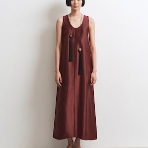 May include: A sleeveless, floor-length dress in a deep burgundy, shimmering fabric with a V-neckline. The dress is accessorized with a long, thin black cord necklace featuring two black tassels and metallic accents. Red thong sandals are worn.