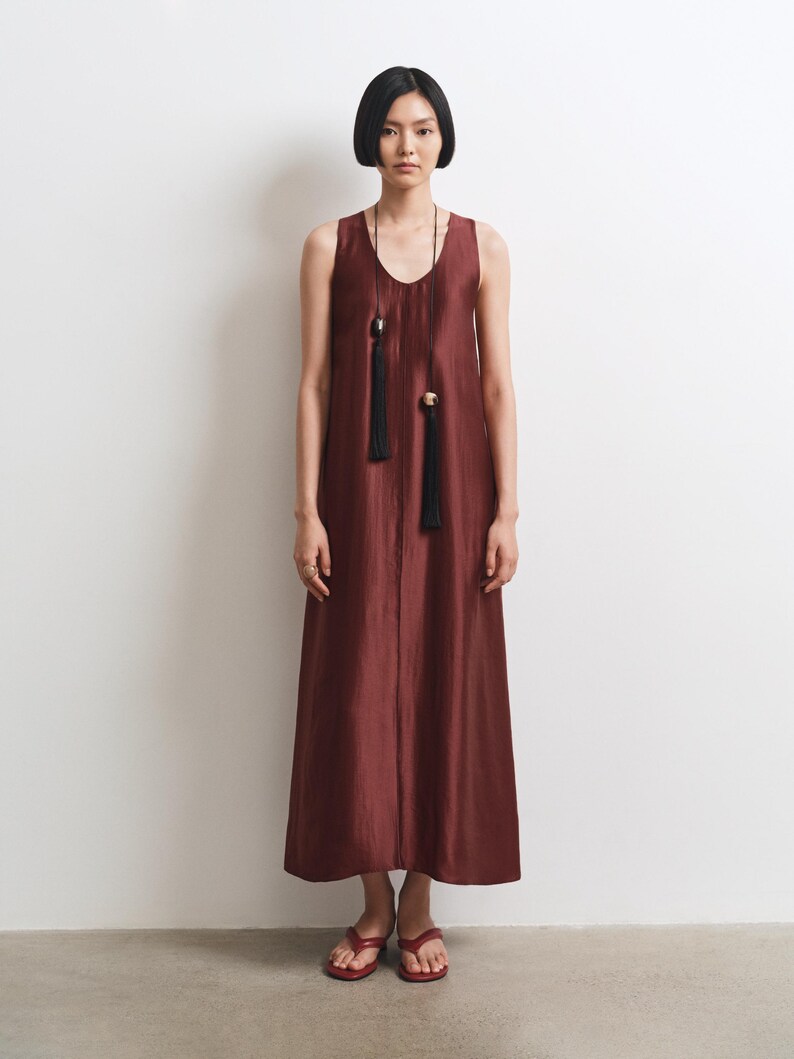 May include: A sleeveless, floor-length dress in a deep burgundy, shimmering fabric with a V-neckline. The dress is accessorized with a long, thin black cord necklace featuring two black tassels and metallic accents. Red thong sandals are worn.